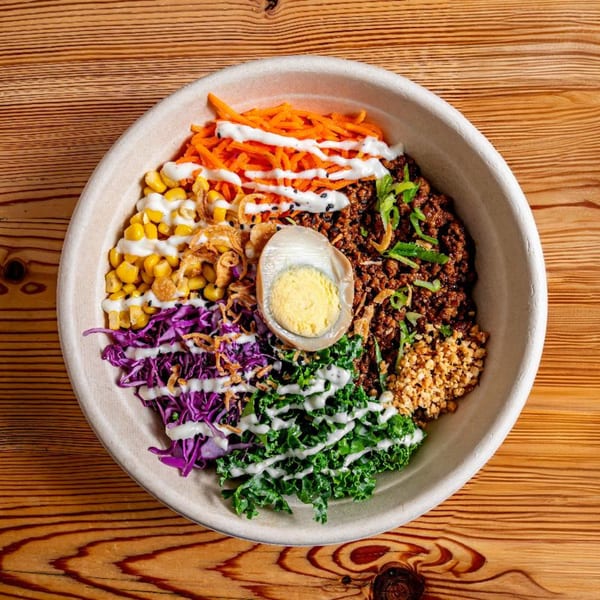 Colorful Asian-fusion bowl with ground meat, egg, corn, carrots, kale, and purple cabbage on wood.