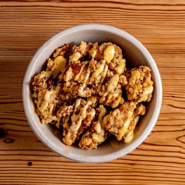 Crispy fried chicken pieces drizzled with a creamy yellow sauce in a light gray bowl on a wooden table.