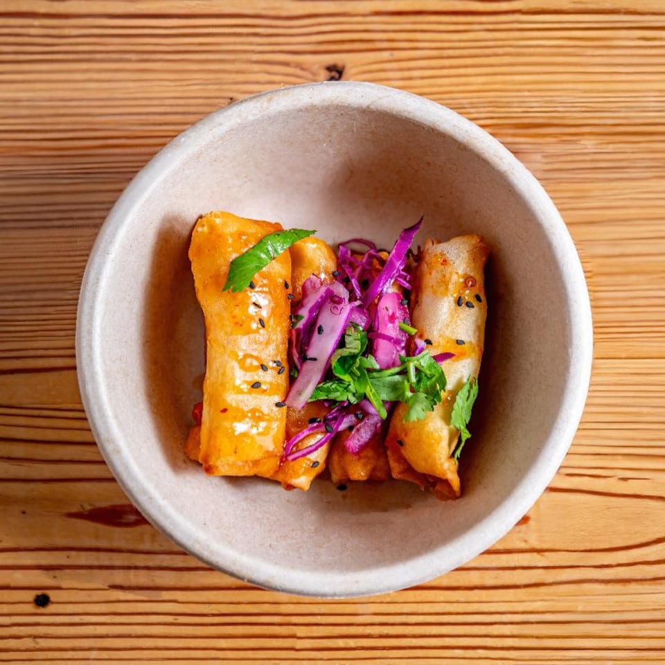 Three golden-fried spring rolls topped with purple cabbage, cilantro, and black sesame seeds in a bowl.