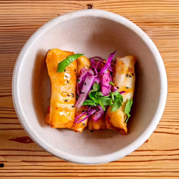 Three golden-brown spring rolls with red cabbage and cilantro in a white bowl on a wooden table.
