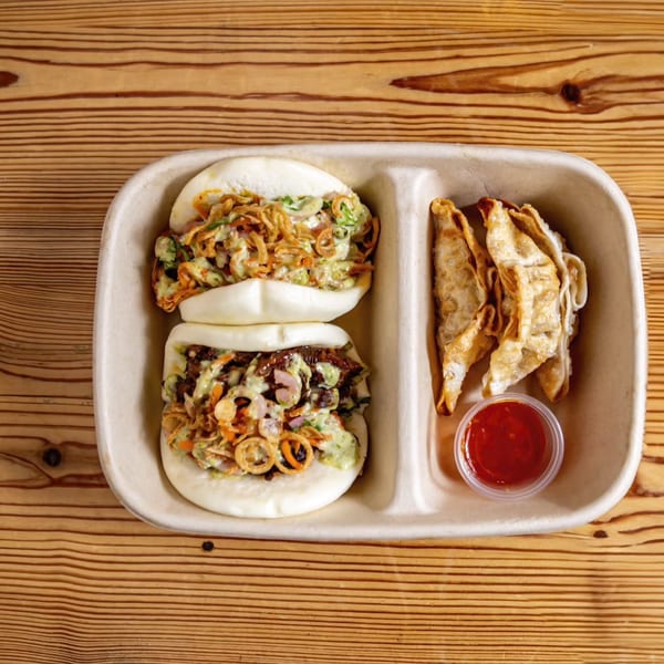 Two bao buns and several fried dumplings with a side of red sauce in a takeout container.