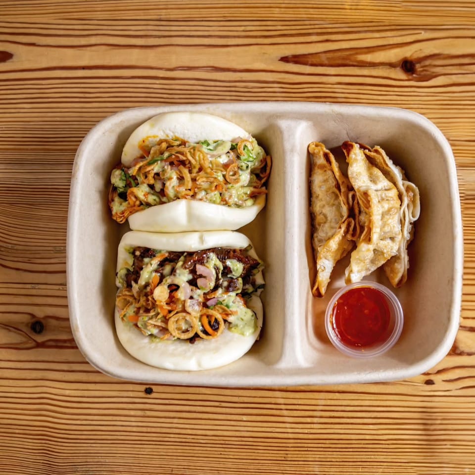 Two bao buns and several fried dumplings with a side of red sauce in a takeout container.