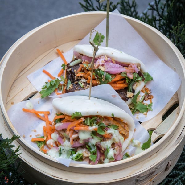 Two Kaya bao buns with meat, pickled onions, carrots, and cilantro in a bamboo steamer.