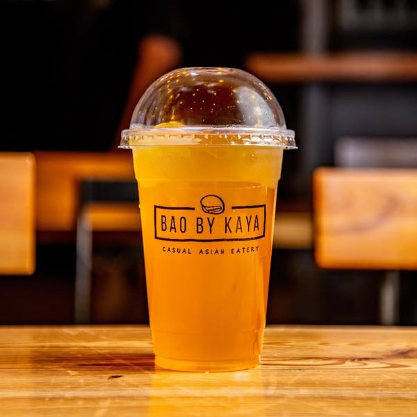 A clear plastic cup of orange-colored drink with the "Bao by Kaya" logo sits on a wooden table.